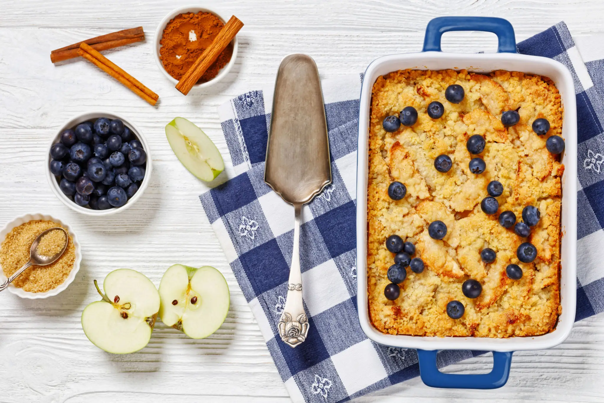 An apple and blueberry crisp in a baking dish, with fresh apples, blueberries and cinnamon quills around it