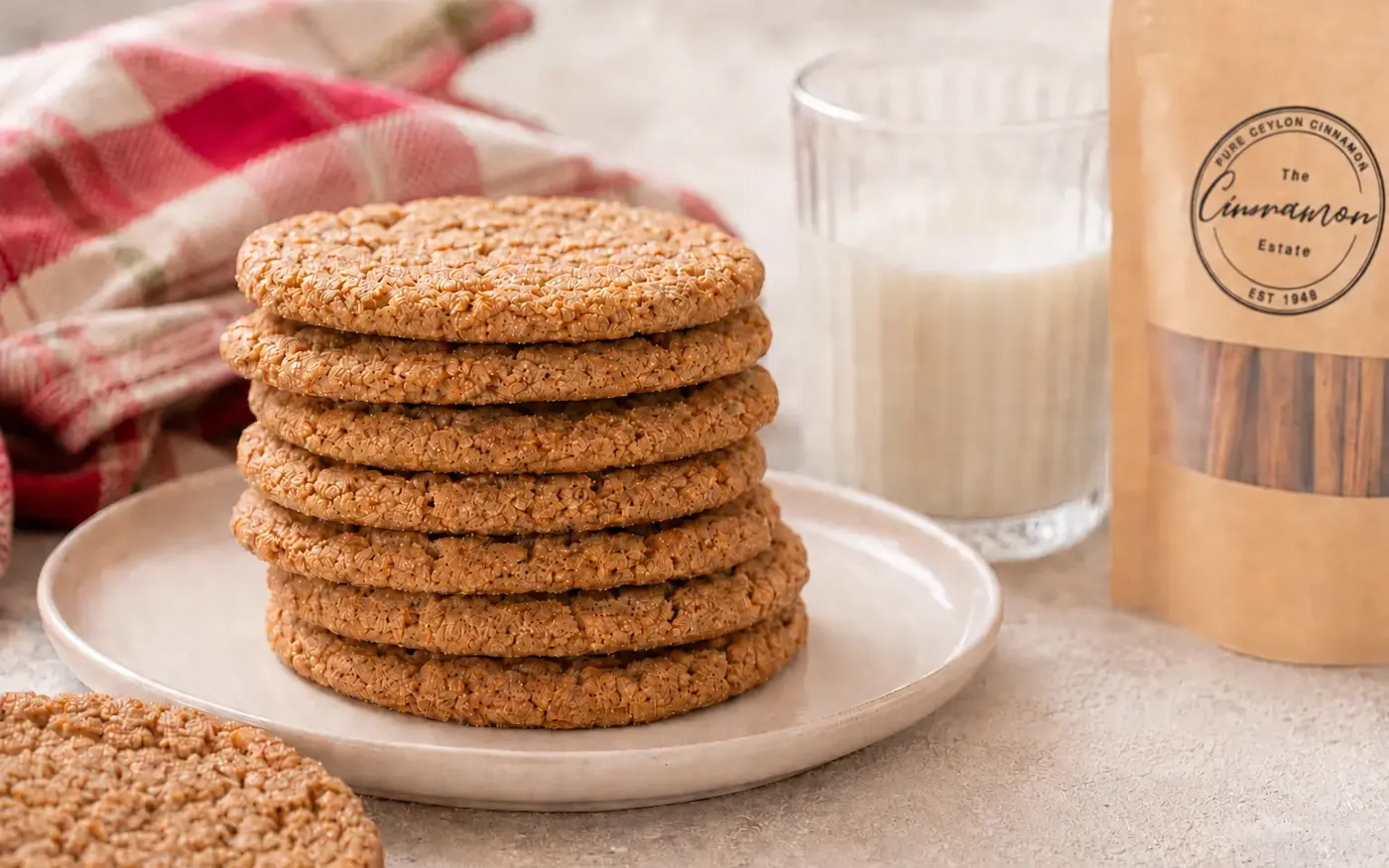 A stack of crackled cinnamon sugar biscuits on a plate with a glass of milk, beside a pouch of The Cinnamon Estate Ceylon cinnamon quills